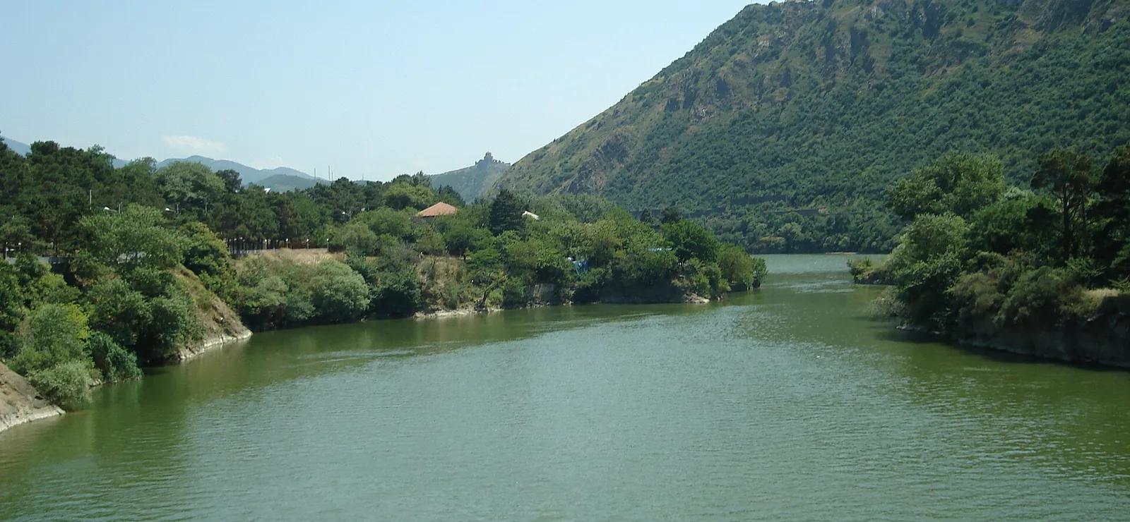 The Aragvi and the Mtkvari meeting below Mtskheta, seen from the water. Green river between wooded banks, with Jvari monastery small on the ridge above.