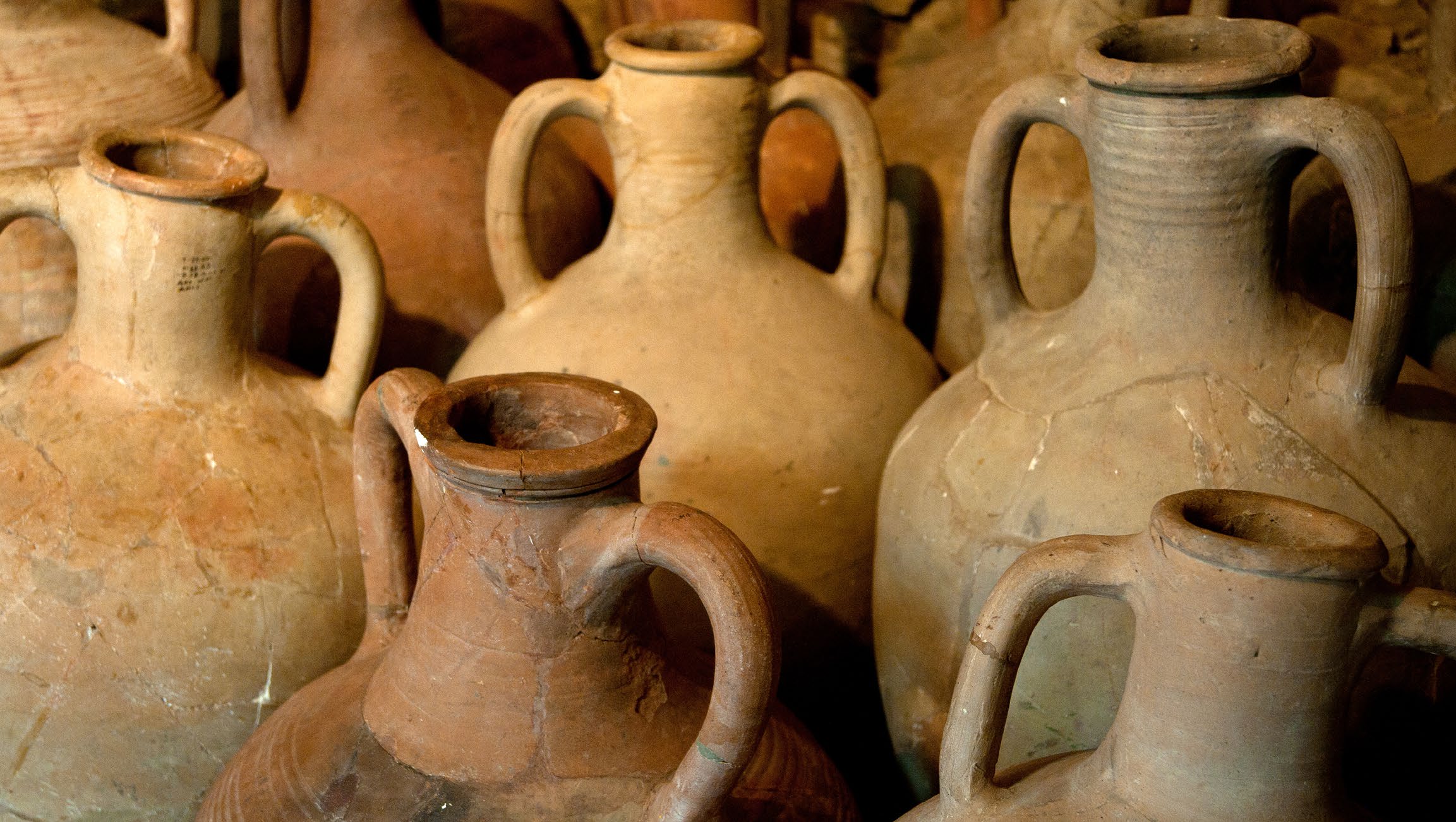 A dense group of terracotta qvevri, cream through rust, cracks and repairs visible