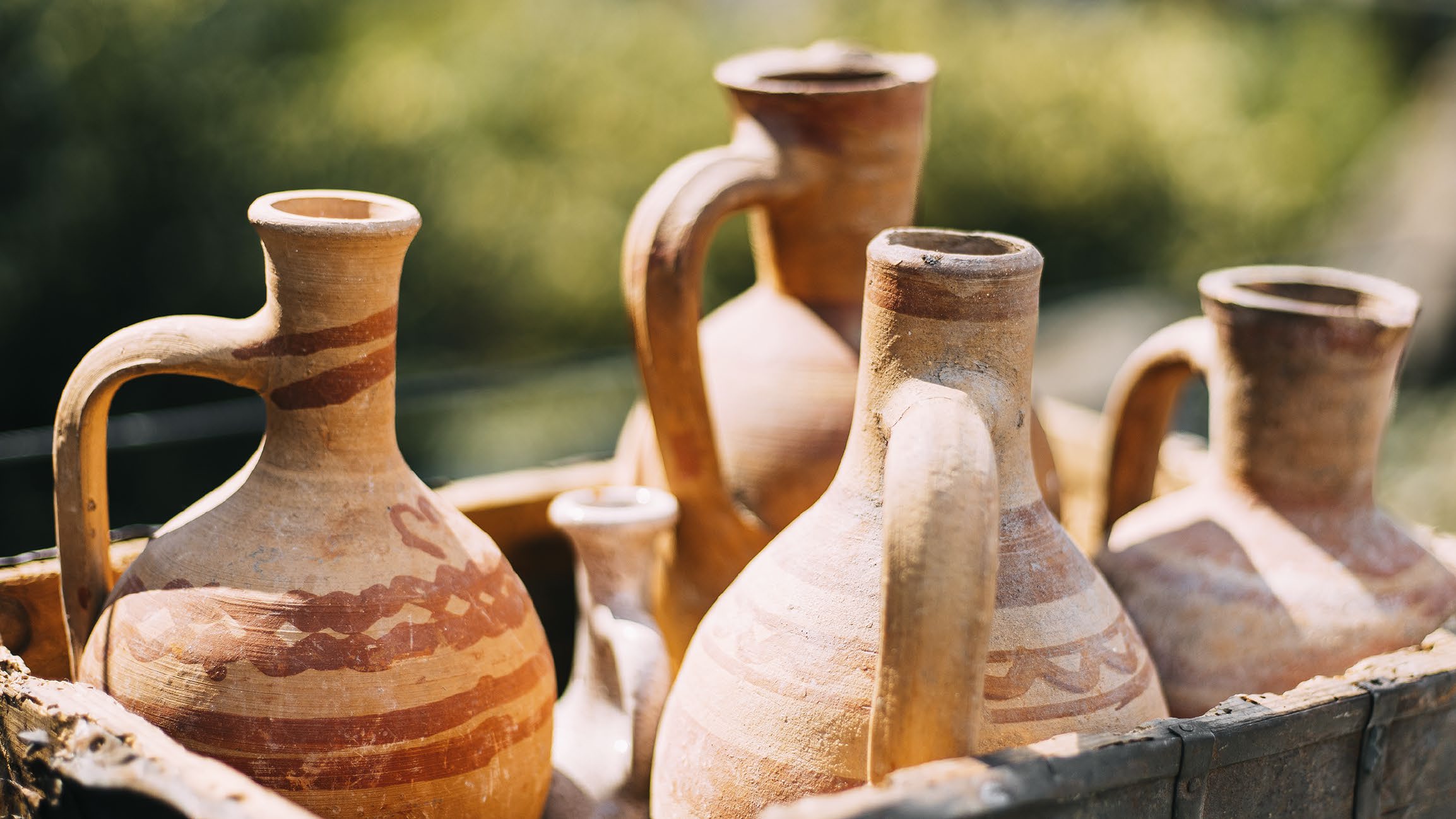 Clay jugs in a weathered wooden crate outdoors in hard sunlight, painted rust red bands and a spiral on the front jug