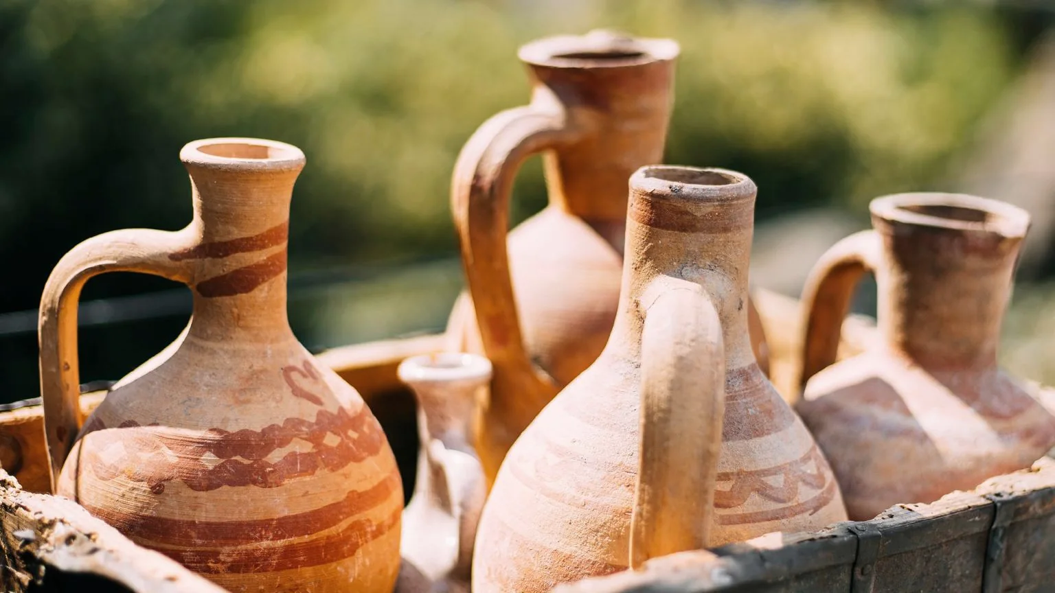 Painted clay jugs standing together in a wooden crate outdoors, green garden light behind them