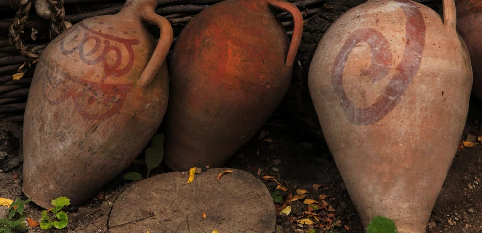 Three large clay qvevri resting against a woven branch fence, the right hand vessel carrying the painted ARAGVI spiral